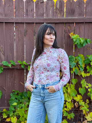 Woman wearing a mesh floral long-sleeve top and jeans standing in front of a wooden fence with greenery.