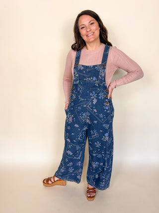 A woman modeling a pair of blue floral print denim overall with a pink ribbed long-sleeve top and brown sandals, standing against a plain beige studio background.