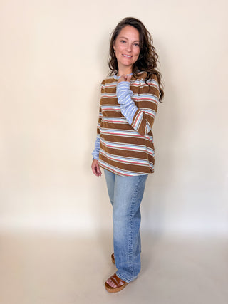 A woman modeling a brown, white, and light blue striped long-sleeve top with light-wash blue jeans and brown slide sandals, standing against a plain beige studio backdrop.