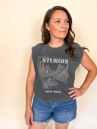 A woman modeling a dark gray sleeveless graphic t-shirt with a white 'meet me inSturgis South Dakota' mountain and motorcycle print, paired with denim shorts against a plain beige background.