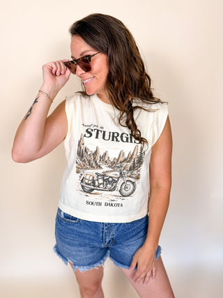 A woman wearing a sleeveless cream-colored graphic t-shirt with a vintage motorcycle and 'STURGIS SOUTH DAKOTA' print, paired with denim shorts, posing against a plain beige background.