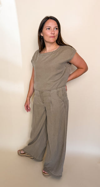 A woman modeling a two-piece linen set consisting of a short-sleeve crewneck top and matching wide-leg pants in a muted taupe color, standing against a plain off-white wall.