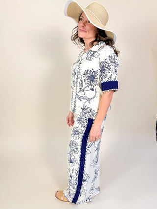 Woman wearing a white dress with blue floral pattern and a wide-brimmed hat on a plain background