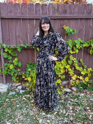 Woman in a black floral dress standing in front of a wooden fence with greenery.