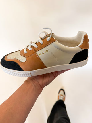 A person holds up a low-top sneaker with a color-blocked design of tan, cream, and black, featuring white laces and a white sole. The brand name 'EST.