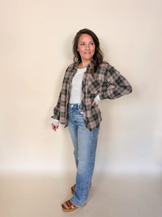 A woman modeling a brown and beige plaid flannel shirt over a white ribbed top, paired with light blue jeans and brown sandals, standing against a plain beige studio backdrop.