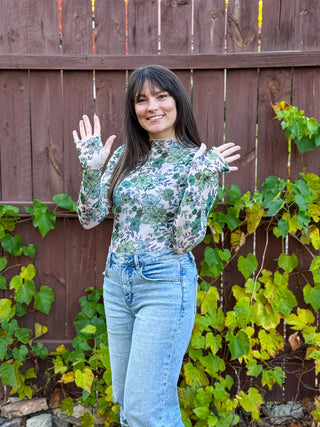 Woman wearing a mesh floral top and jeans standing in front of a wooden fence with greenery.