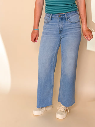 A person wearing light blue wide-leg denim jeans, a green striped top, and white sneakers, standing against a warm beige studio background.