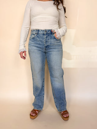 A model wearing a pair of light-wash blue straight-leg jeans, a white ribbed long-sleeve top with lace trim, and brown platform sandals, standing against a plain beige studio backdrop.