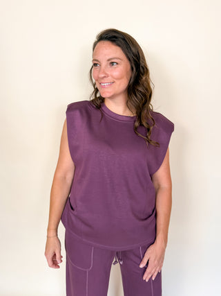 A woman modeling a sleeveless crewneck top and matching drawstring pants in a solid purple color, standing against a plain off-white background.