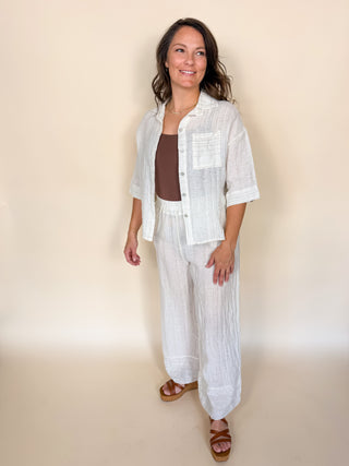 A woman modeling a white textured linen two-piece set consisting of a short-sleeve button-up shirt and matching wide-leg pants, worn over a brown tank top and brown sandals, against a solid beige studio background.