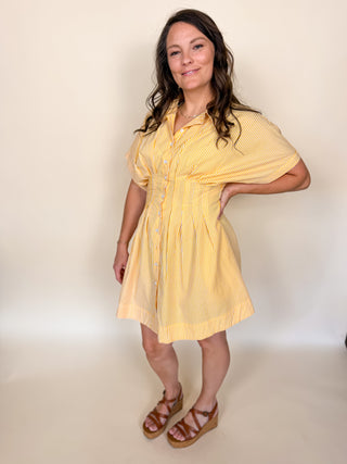 A woman modeling a short-sleeve yellow and white striped button-up shirt dress with a gathered waist, paired with brown strappy sandals, against a plain beige studio backdrop.