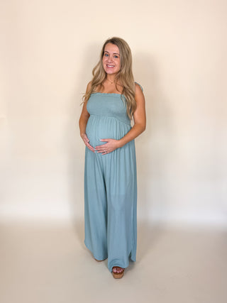 A pregnant woman with blonde hair stands against a plain beige studio background wearing a dusty blue sleeveless maternity jumpsuit.
