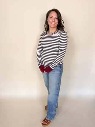 A woman modeling a long-sleeve crewneck top with horizontal black and white stripes, paired with light blue jeans and brown slide sandals, standing against a plain beige studio background.