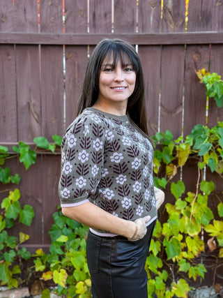 Woman wearing an olive short sleeve patterned sweater with lavender floral details standing in front of a wooden fence with greenery.