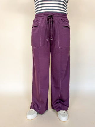 A person wearing wide-leg purple sweatpants with a drawstring waist and side pockets, paired with a black and white striped top and white sneakers, shown against a plain beige studio background.