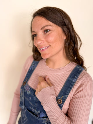 A woman with brown hair wearing a pink ribbed long-sleeve top and blue floral overalls, posing against a plain cream-colored wall.