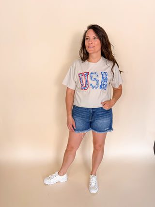 Woman wearing a 'USA' t-shirt and denim shorts on a beige background