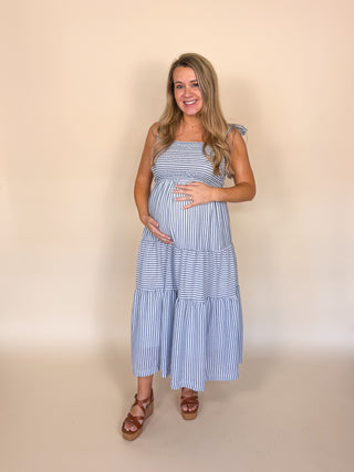 A pregnant woman wearing a blue and white striped tiered maxi dress with shoulder tie details, standing against a solid beige studio background.