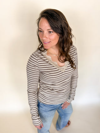 A woman wearing a beige and brown horizontally striped long-sleeve top with a lace-trimmed scoop neckline, paired with light blue jeans, photographed against a plain cream studio background.