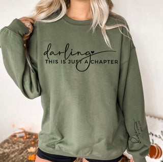 Graphic crew sweatshirt reading “Darling This Is Just a Chapter” with sleeve text “Not the Whole Story” available in blush pink, olive green, and cream.