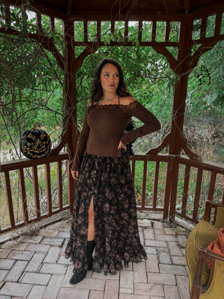 A woman stands in a wooden gazebo wearing a brown off-the-shoulder long-sleeve top and a long black floral skirt with a thigh-high slit, paired with black boots. Trees and outdoor scenery are visible in the background.